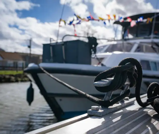 Gros plan d'un cordage noir noué à une bitte d'amarrage dans le port fluvial d'Aire-sur-la-Lys, avec des bateaux de plaisance amarrés en arrière-plan par beau temps