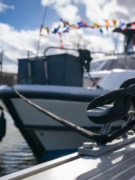 Gros plan d'un cordage noir noué à une bitte d'amarrage dans le port fluvial d'Aire-sur-la-Lys, avec des bateaux de plaisance amarrés en arrière-plan par beau temps
